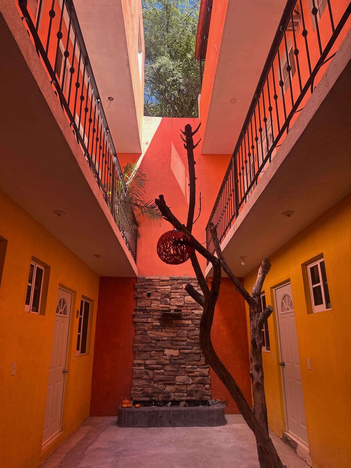 El patio de Hostal Villegas: muros naranja y amarillo, barandales de hierro en el piso de arriba, un tronco seco con una lámpara de mimbre y una fuente de piedra al fondo